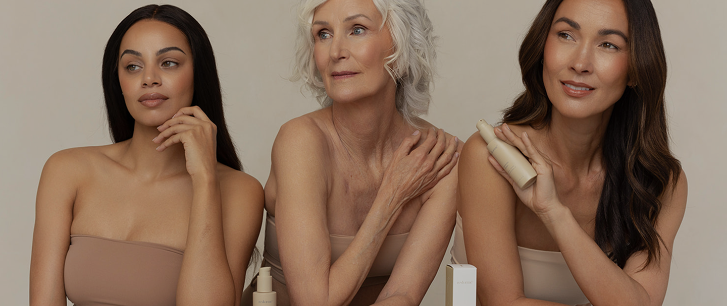 Three women of different ages and ethnicities posing together against a warm beige background, wearing matching beige strapless tops and holding cosmetic bottles.